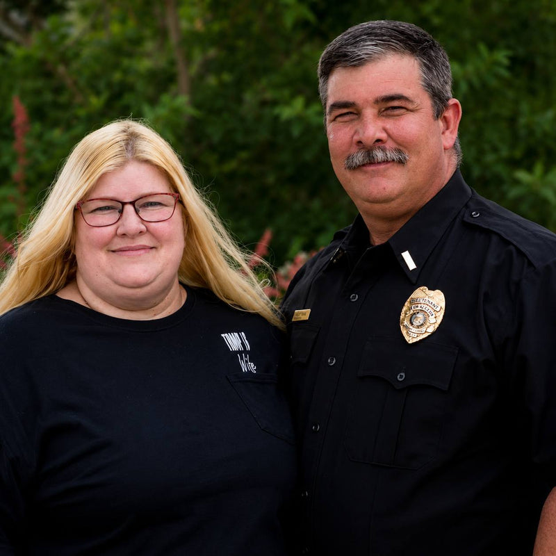 Two individuals, one in a black shirt with a logo and the other in a police uniform with a badge, standing outdoors.