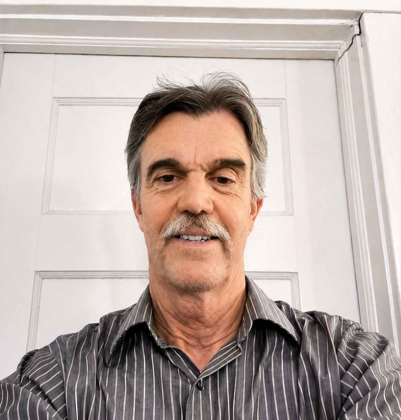 Man with a mustache wearing a striped shirt in front of a white door.