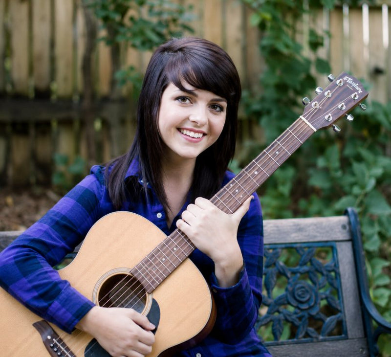 Young woman smiling while holding an acoustic guitar outdoors on a bench, wearing a blue plaid shirt with a wooden fence and greenery in the background.