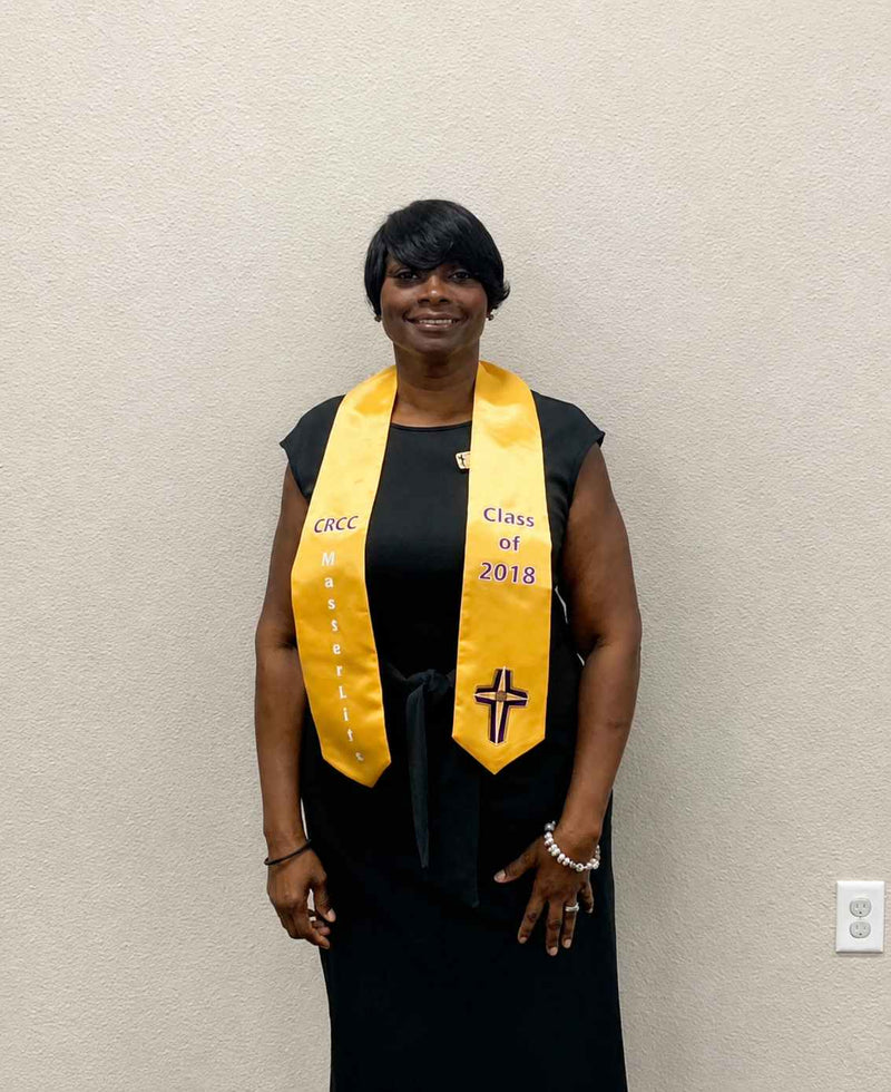 Person wearing a black graduation gown with a yellow stole against a beige wall.